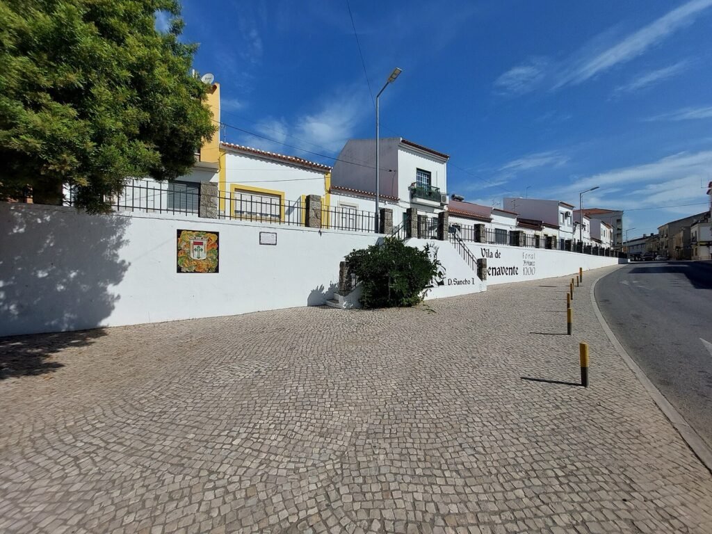 Uma estrada a curvar junto a um muro branco e a uma fila de casas, com passeio em calçada em primeiro plano.