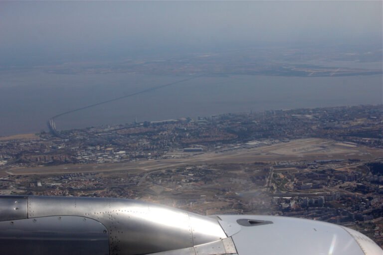 Aerial photo of Lisbon's airport runways surrounded by city districts, with a long bridge crossing a wide estuary in the background.