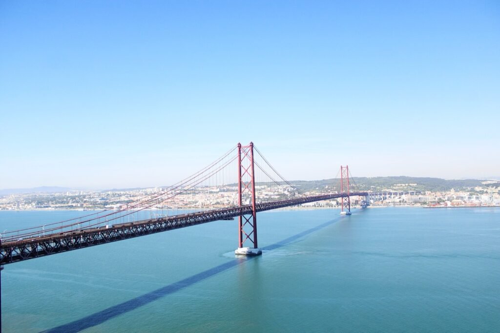 Red suspension bridge seen from the south bank, with Lisbon behind it.