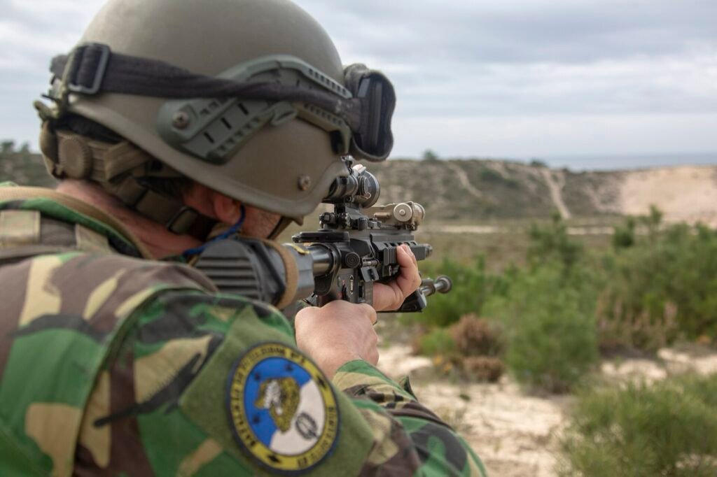 A soldier aims a rifle across sandy scrubland with pine trees.